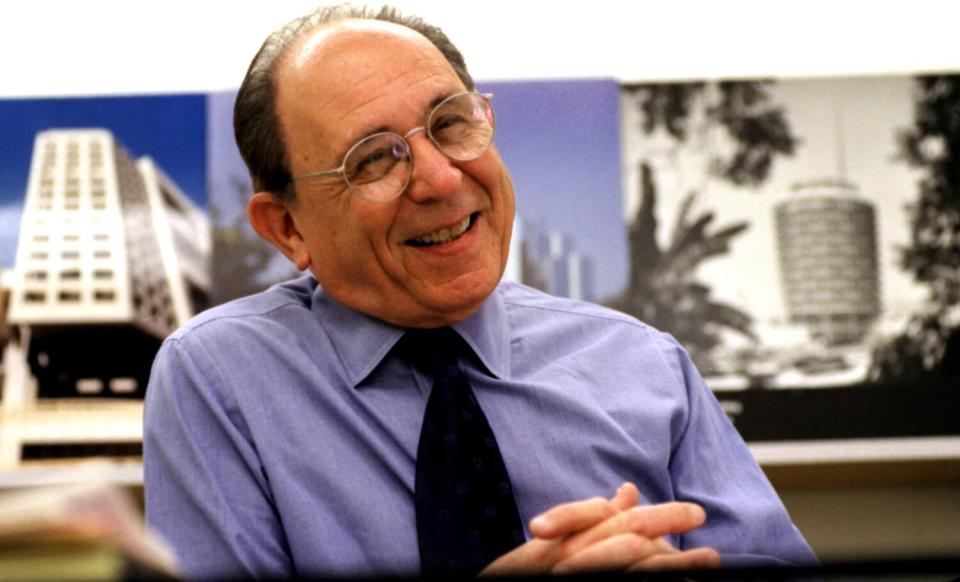 A man in a blue shirt and black tie smiles with clasped hands, with photos of buildings behind him