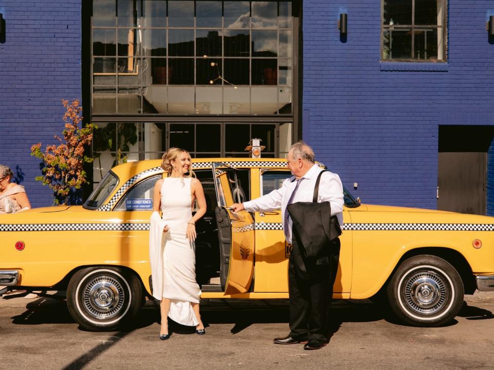 A bride steps out of a yellow taxi as a man opens the door for her.