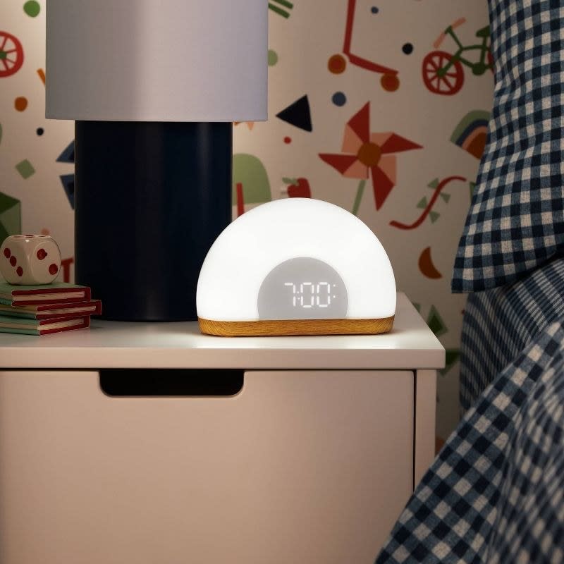 Modern digital alarm clock on a nightstand displaying 7:00, with books and a lamp nearby, for a home decor shopping article