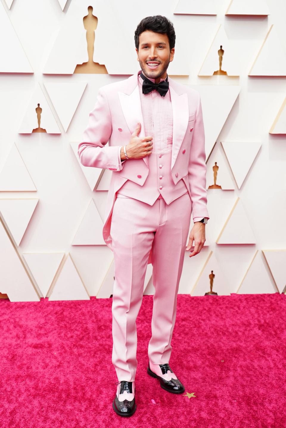 Sebastian in a pink suit with a waistcoat, low vest, shirt with pleating, trousers, and pink and black wing-toed shows. He has a black bow tie on.