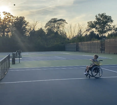 A little boy in a wheelchair swings a yellow racket on a tennis court as the sun sets. (Courtesy of Jason and Keely Roberts)