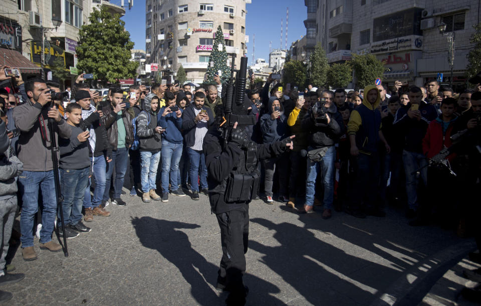 Palestinians take pictures with their phones of a masked armed Palestinian carry weapons during a celebration marking the 55th anniversary of the Fatah movement, in the West Bank city of Ramallah, Tuesday, Dec. 31, 2019. (AP Photo/Majdi Mohammed)