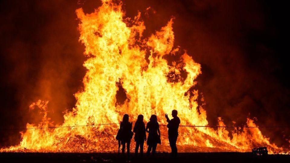 Silhouettes of people standing in front of a bonfire set a lit in orange, yellow and red flames 