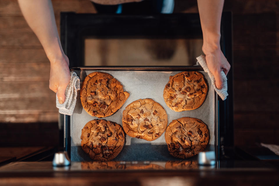 A person takes a tray of five freshly baked cookies out of the oven using a pair of cloth towels