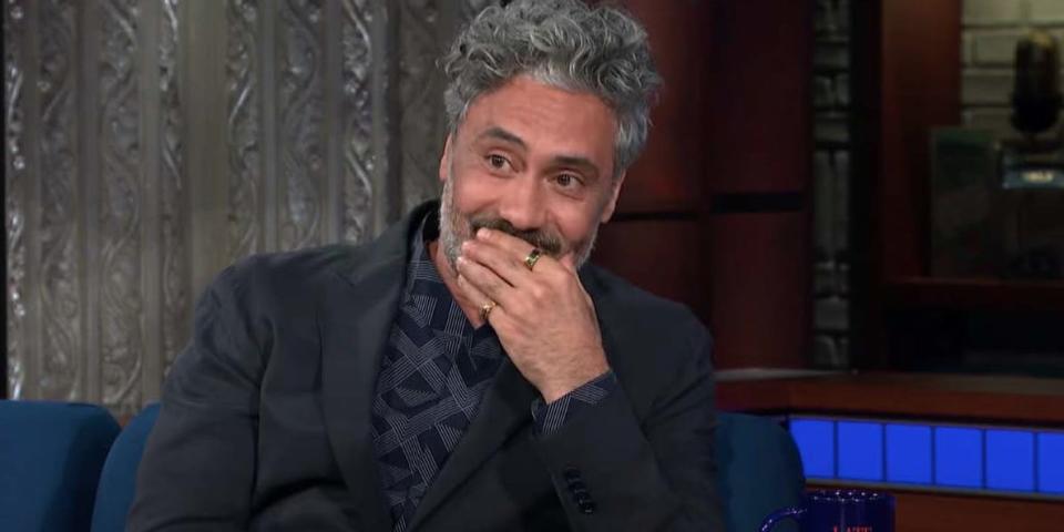 Taika Waititi putting his hand to his mouth while sitting a chair