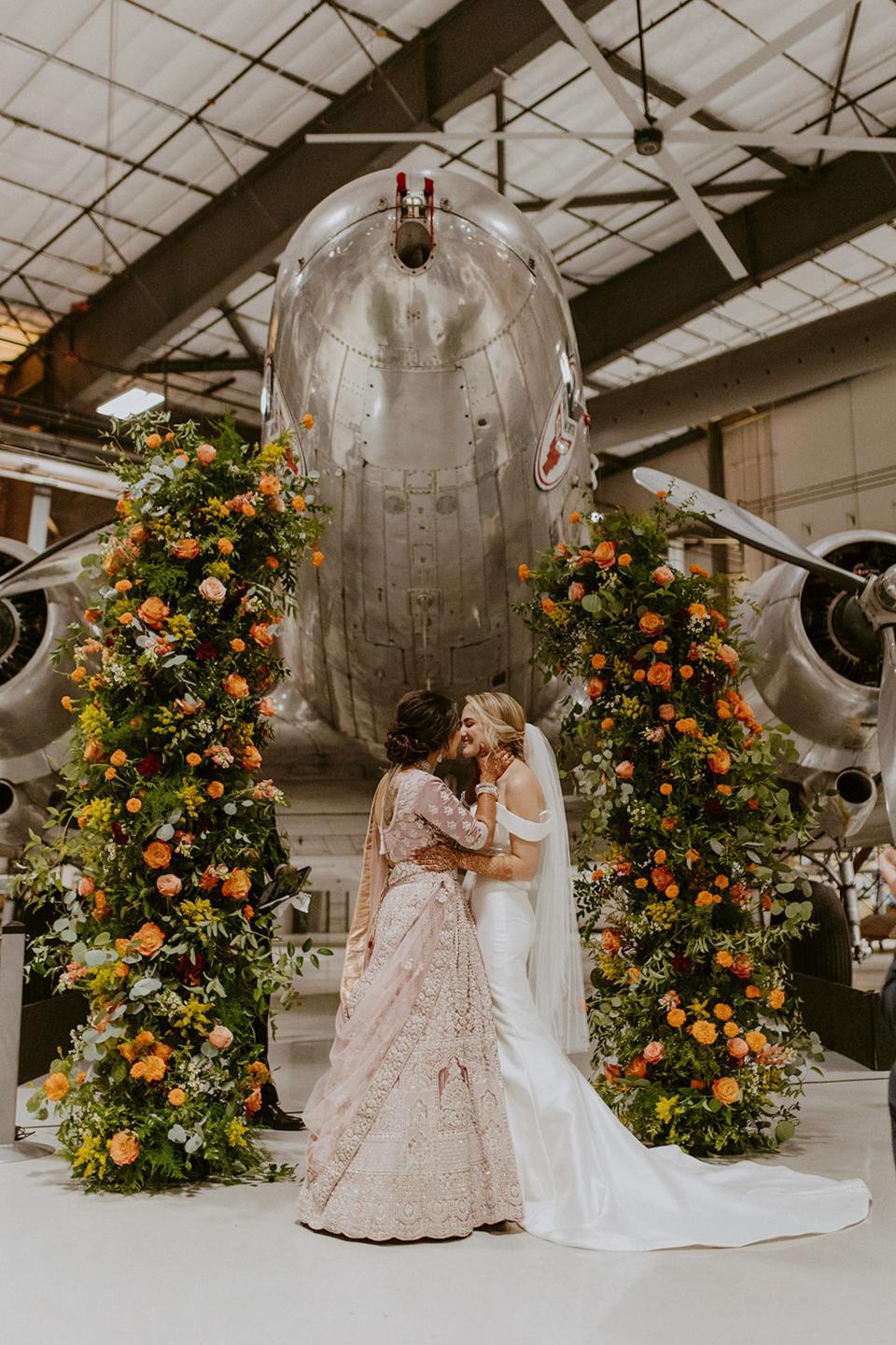 Two brides kiss at their wedding altar surrounded by a floral archway in front of a plane.