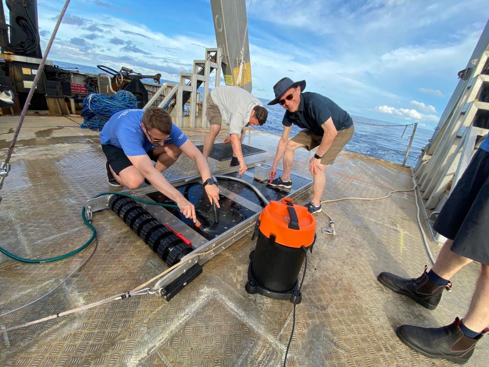 Loeb smiles as he holds a vacuum to collect material from a sled. Other colleagues are cleaning the sled as well. The team are onboard a boat.