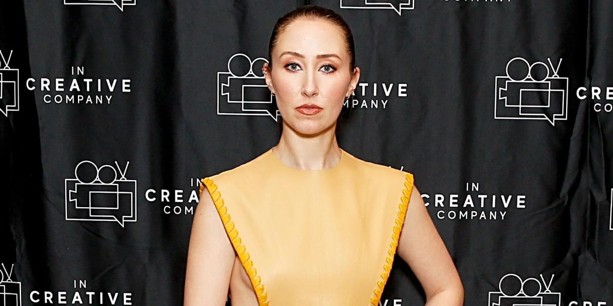 erin doherty, standing with left hand on hip looking at the camera, wearing a yellow dress, dark hair pulled back in a bun