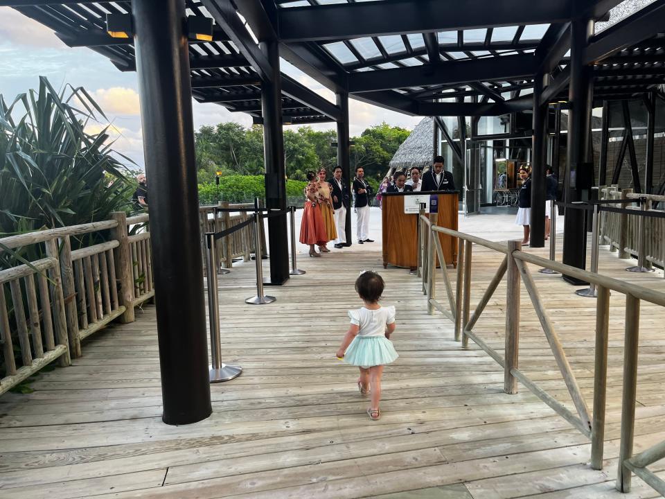 The writer's daughter, wearing a blue skirt and white shirt, walks on a wooden bridge toward staff members in suits and women in red and orange skirts