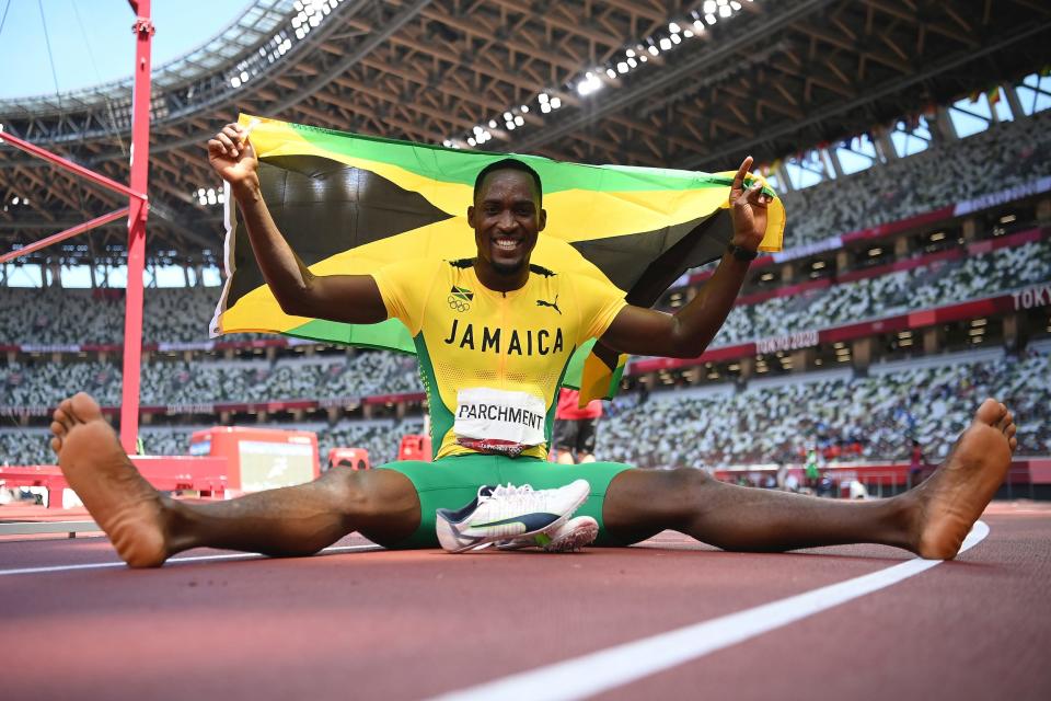 Hansle Parchment reacts to winning gold at the Tokyo Olympics, sitting on the track with his shoes off and a Jamaican flag held over his head