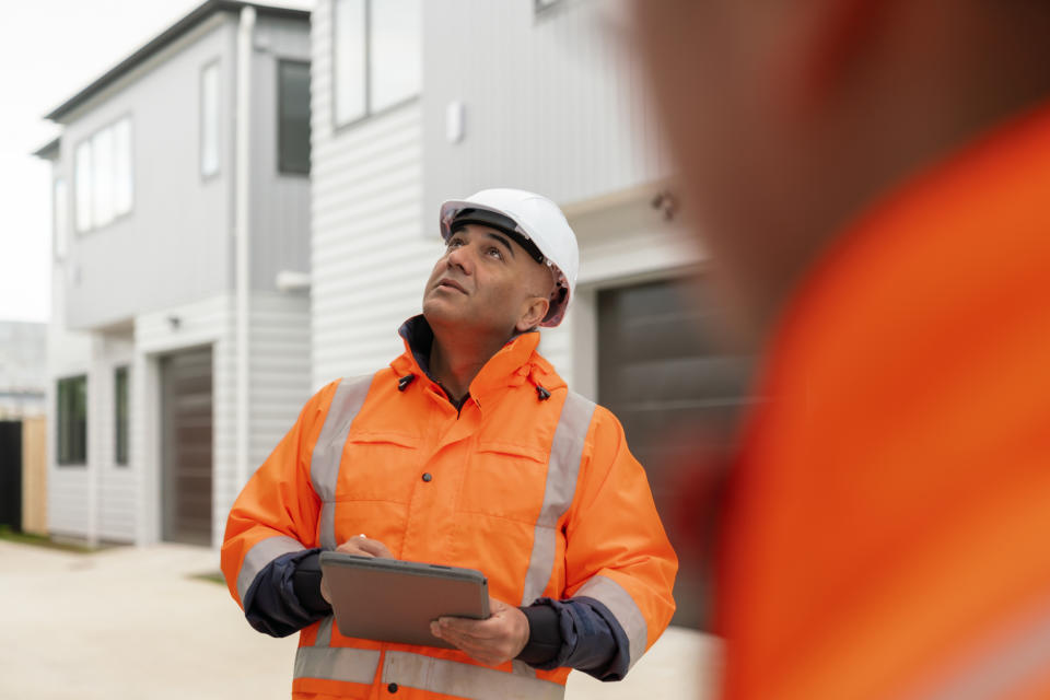 A construction worker wearing a hard hat and an orange safety jacket examines the exterior of a building, holding a clipboard. Another worker is partially visible