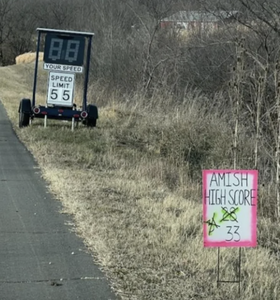 "Speed limit 55" road sign with digital "your speed" above, and handwritten sign ahead of it: Amish high score 33, with 28 and 31 crossed out