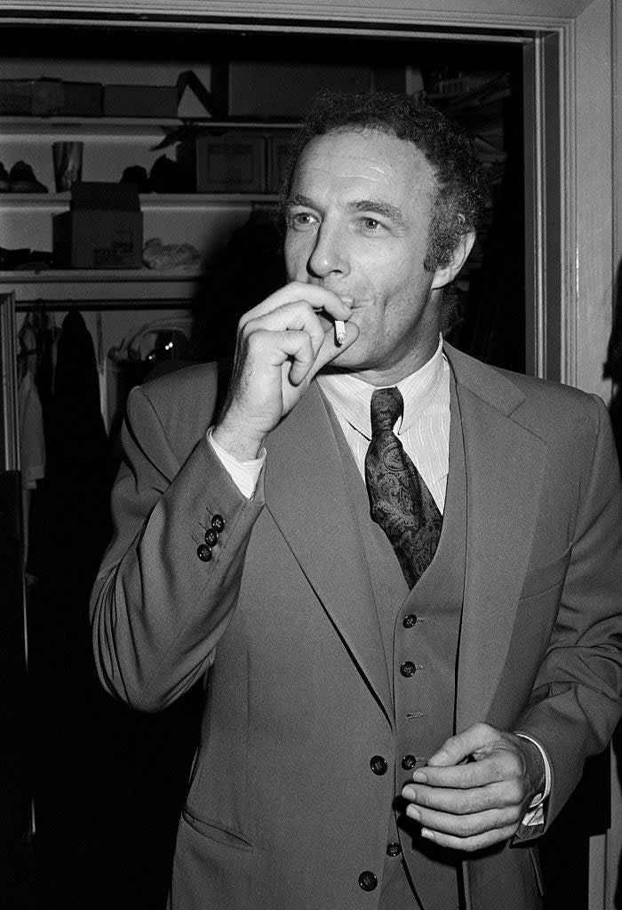 James Caan, wearing a three-piece suit with a patterned tie, is standing indoors, smoking a cigarette and looking off to the side