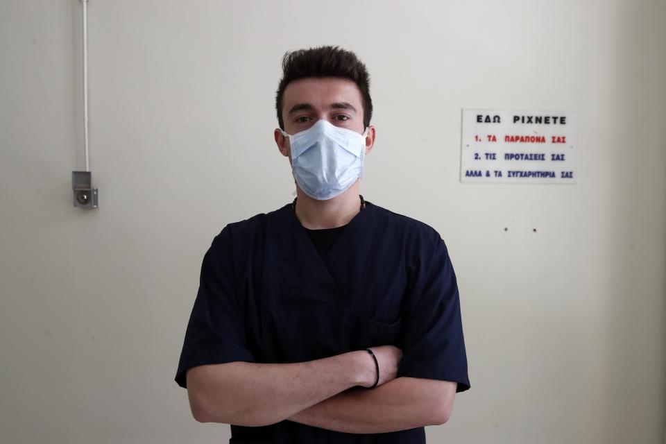 In this photo taken on Friday, May 8, 2020, medical student Nikolaos Katsanakis poses for a photo at the entrance of the COVID-19 Clinic at Sotiria Hospital in Athens. Greece's main hospital for the treatment of COVID-19 is also the focus of a hands-on training program for dozens of medical students who volunteered to relieve hard-pressed doctors from their simpler duties while gaining a close peek at the front lines of a struggle unmatched in modern medical history. The sign below, placed above a box that has since been removed, reads ''This is where you leave your complaints, your suggestions and your congratulations too.'' (AP Photo/Thanassis Stavrakis)