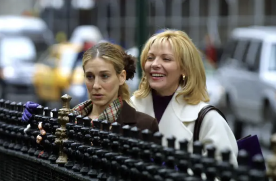 A photo from the set of Sex and the City, showing Carrie and Samantha looking at something while standing behind a park fence