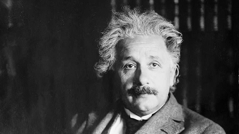 albert einstein sitting in front of a bookcase with his arms folded