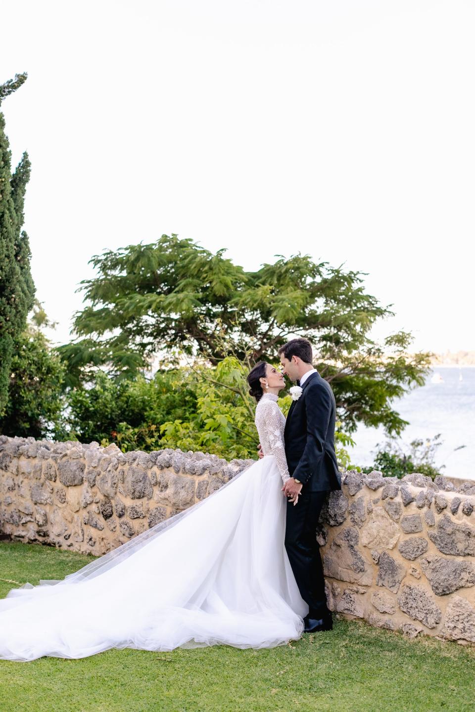 A bride and groom lean towards each other and hold hands in front of a stone wall overlooking the ocean.