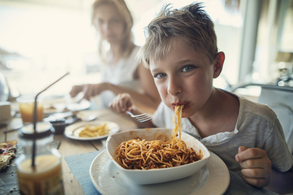 A boy eating pasta in a restaurant. His mother can be seen blurred behind him.