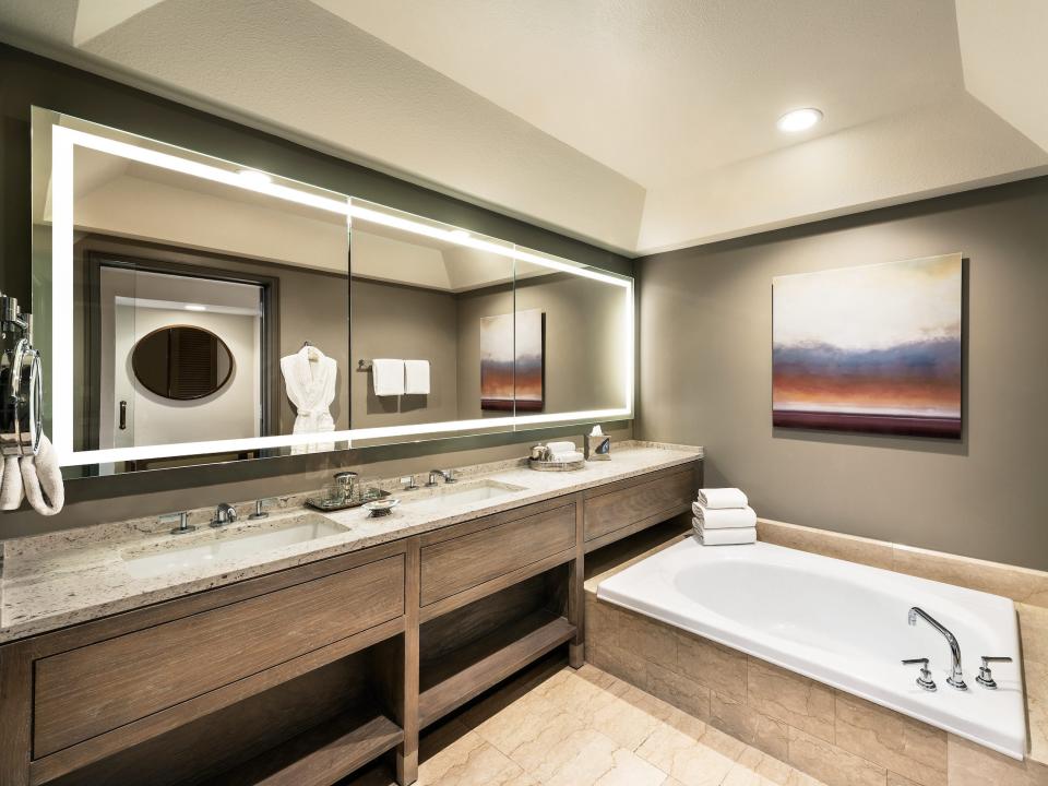 Inside a gray, modern bathroom with a white tub on the right and two sinks and a vanity on the left