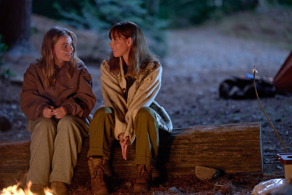 Maisy Stella and Aubrey Plaza sitting on a log on the beach in a movie scene.