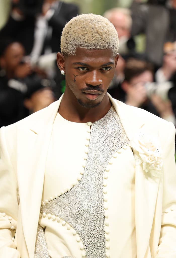 Lil Nas X wearing a embellished suit with an intricate beaded pattern as he attends an event with photographers in the background