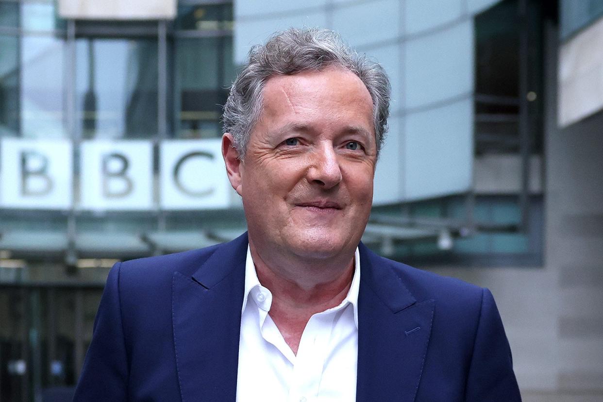 Piers Morgan, in a blue suit jacket and white button-down shirt, stands outside a BBC building and smiles slightly.