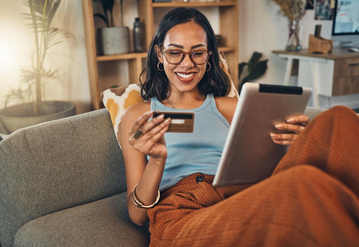 Smiling mixed race woman using credit card for ecommerce on digital tablet at home. Happy hispanic sitting alone on living room sofa, using technology for ebanking. Relaxing, ordering, buying online