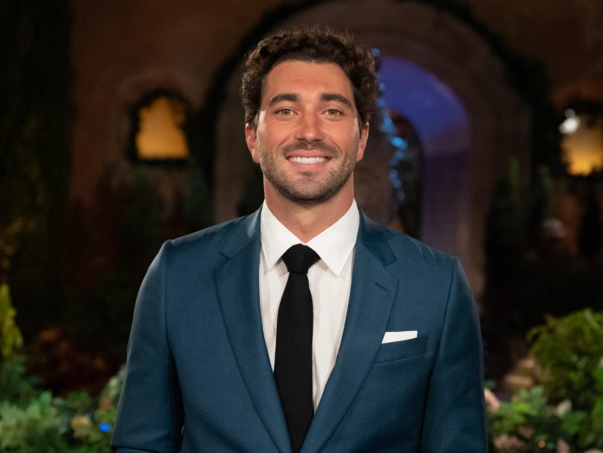 Joey Graziadei smiles in front of the Bachelor mansion in a blue suit, white shirt, and black tie.