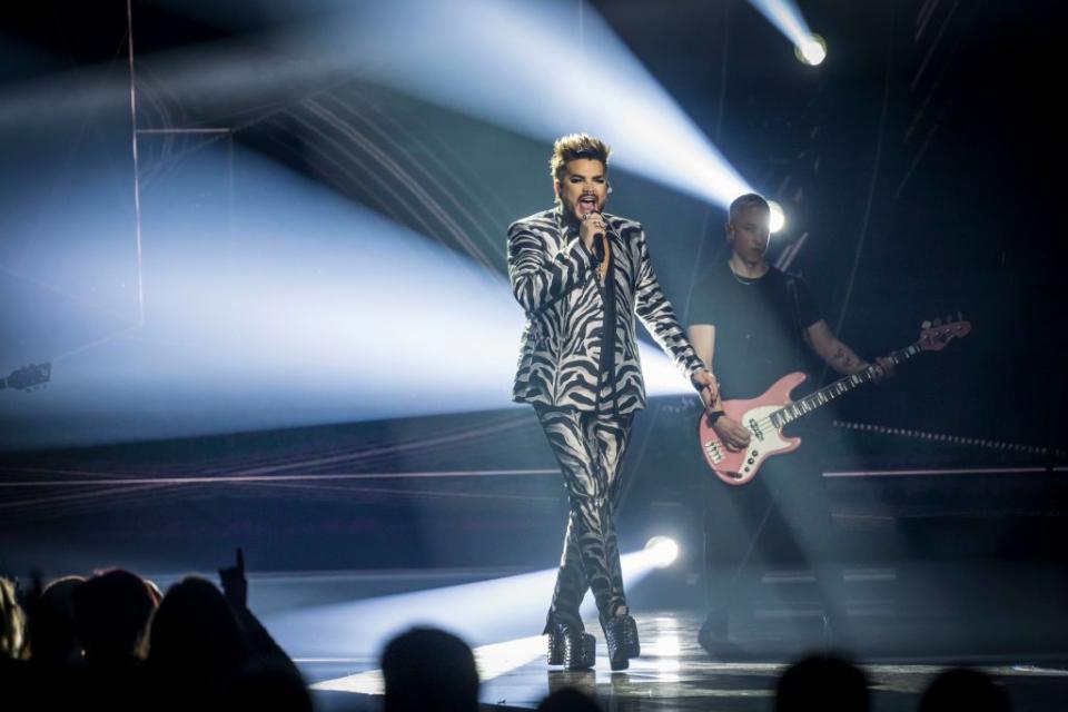 adam lambert sings into a microphone he holds while standing on a stage in a zebra print suit and platform shoes