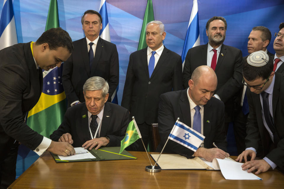 Prime Minister Benjamin Netanyahu, center, and Brazilian President Jair Bolsonaro, second left standing, watch as Mr. Egal Unna, Chief Executive Director of the new Cyber Technologies, seated left, and Mr Agusto Heleno, Minister of State of the Brazilian Institutional Security Cabinet, seated right, sign economic agreements at the prime minister's office in Jerusalem, Sunday, March 31, 2019. (Heidi Levine/Pool Photo via AP)