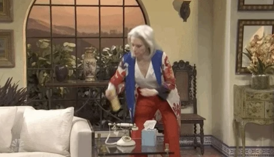 Kate McKinnon, wearing a multicolored cardigan and bright pants, is spraying a phone with disinfectant in a living room set on a show