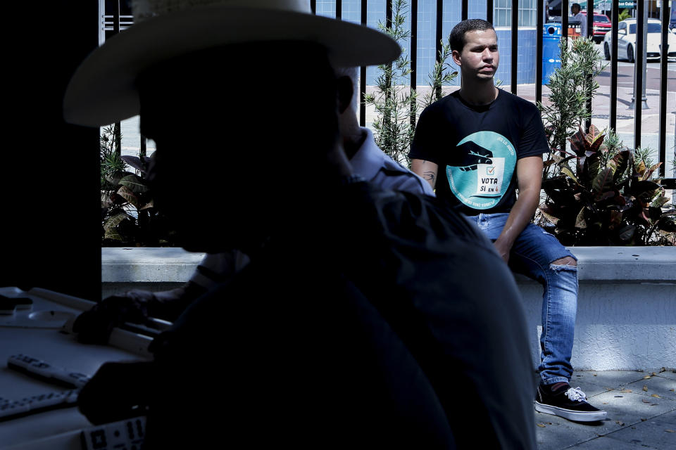 In this Wednesday, June 12, 2019 photo, Andy Vila, right, sits at Domino Park, where elderly men at left play dominoes, in the Little Havana neighborhood of Miami. Although the 21-year old was raised in a politically conservative Cuban American household, he is part of a wave of young Americans who are embracing socialism. His political beliefs have caused deep rifts within his family and social circles. (AP Photo/Ellis Rua)