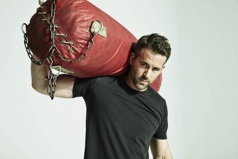 ryan reynolds, ryan reynolds workoutactor, wearing a black t shirt, holding a large, red punching bag on one shoulder