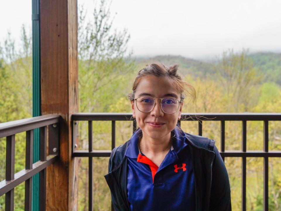 The author in a blue polo standing on the patio with the black fence and mountains and trees in the background. There's a wooden pillar on the left side at the fence's corner. The sky is gray and hazy.