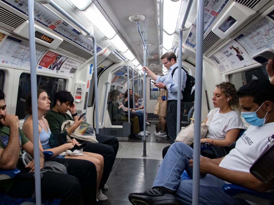 Passengers sit on a London Tube carriage, one standing up holding the Jubilee Line's grey pole.