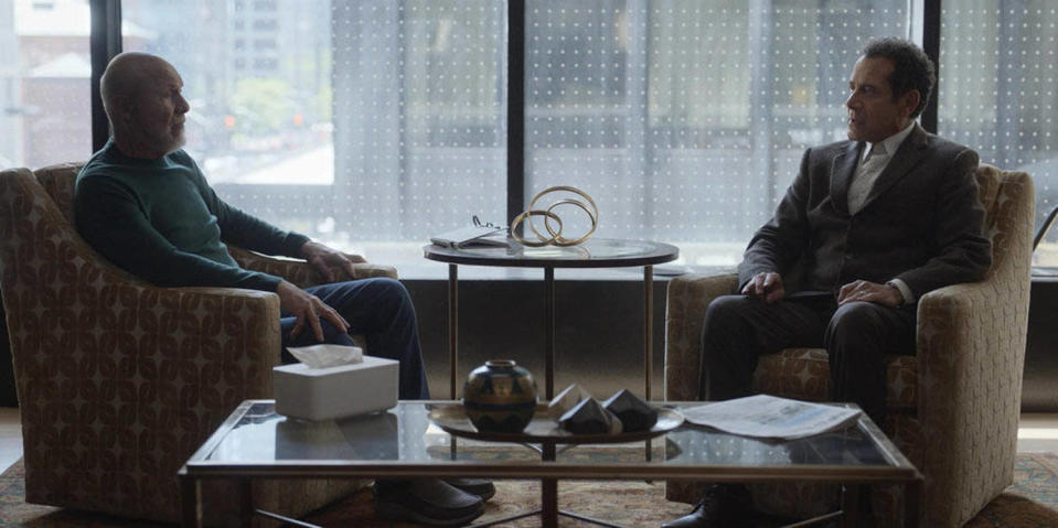 Two men sit in an high-end office in separate chairs looking at each other. One. is in a green T-shirt and jeans, the other a brown suit. (Courtesy of Peacock)