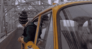 A person wearing a knitted hat leans out of a moving yellow taxi driving over a bridge