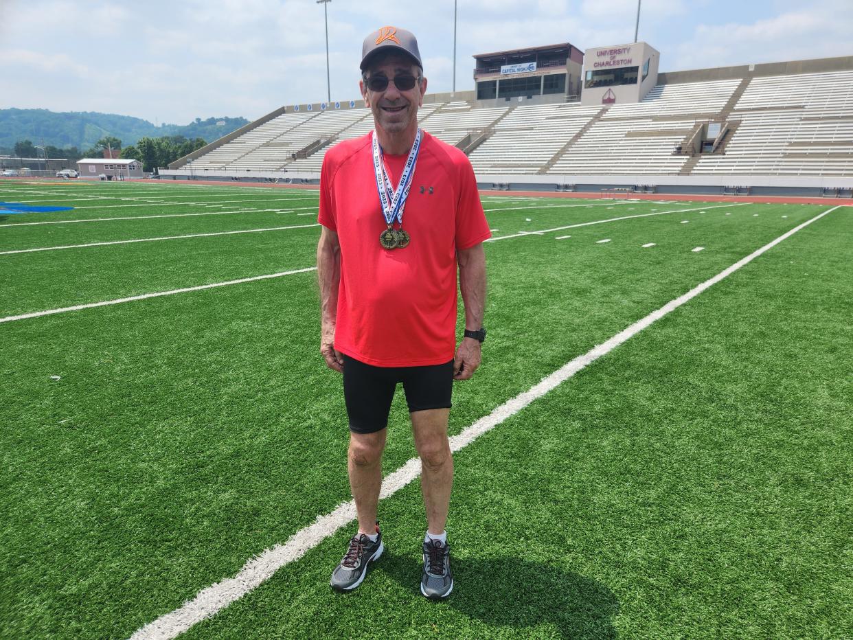 A man in a T-shirt and shorts wearing sports medals standing on an athletic field.