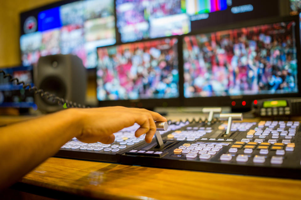 Hands operating a broadcast control panel with multiple monitors showing blurred visuals in a professional studio setting