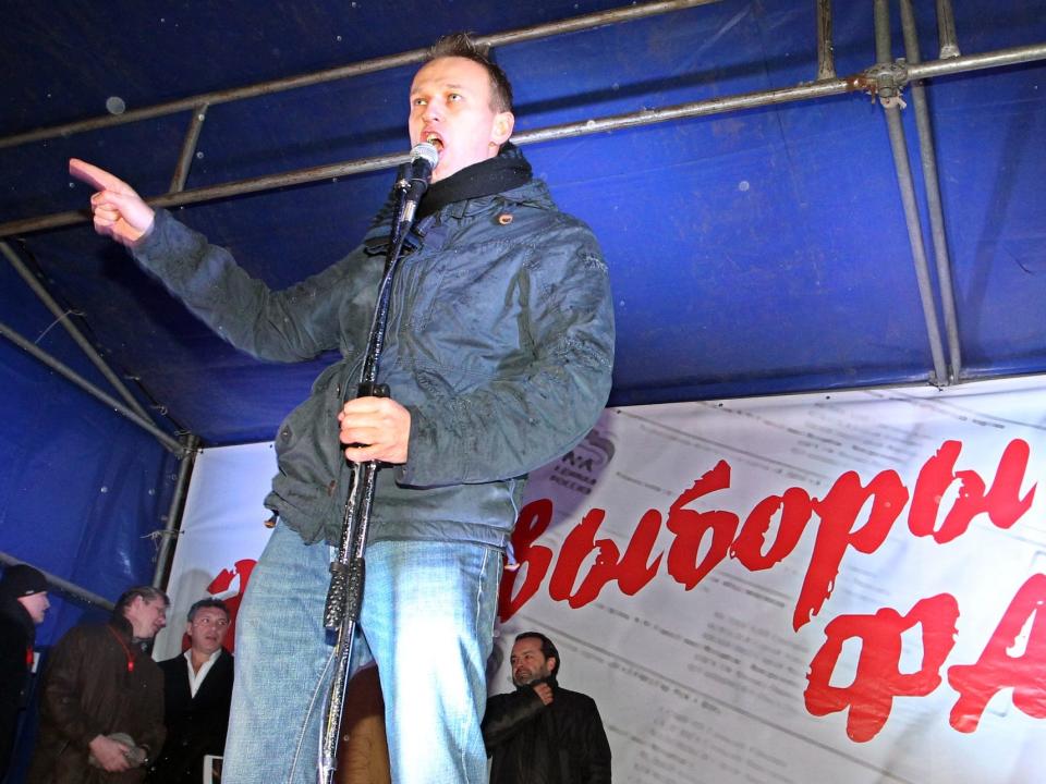 Alexei Navalny speaks into a microphone and points in the distance in front of a sign with words in Russian.