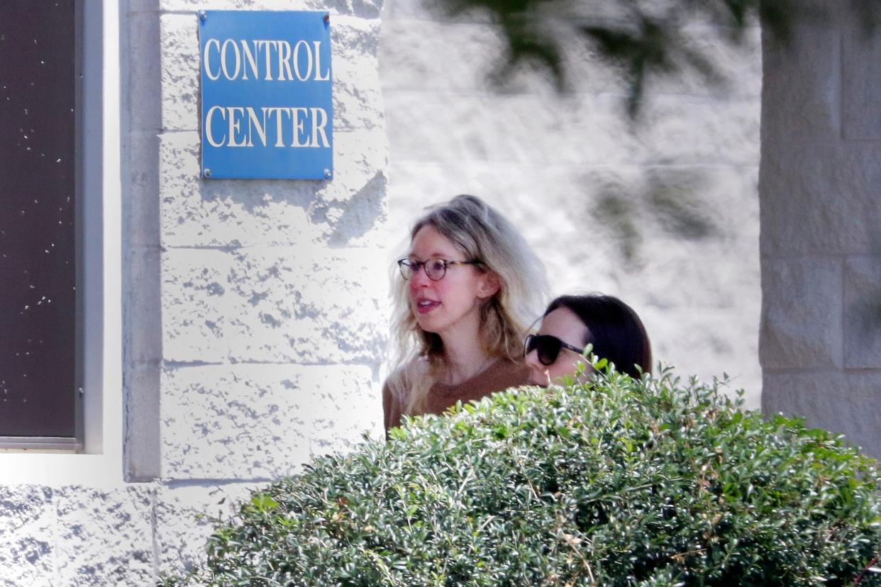 Elizabeth Holmes walking past a bush while heading toward a white-brick building with a blue sign that reads "Control Center" next to a window.