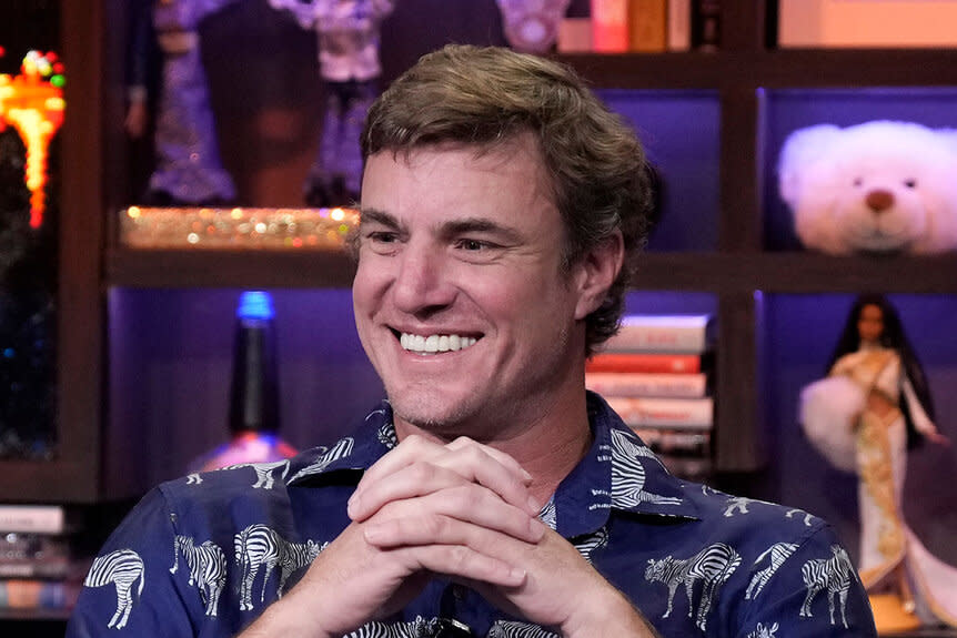 Shep Rose smiling in a navy, zebra patterned, shirt in front of the WWHL step and repeat in New York City.