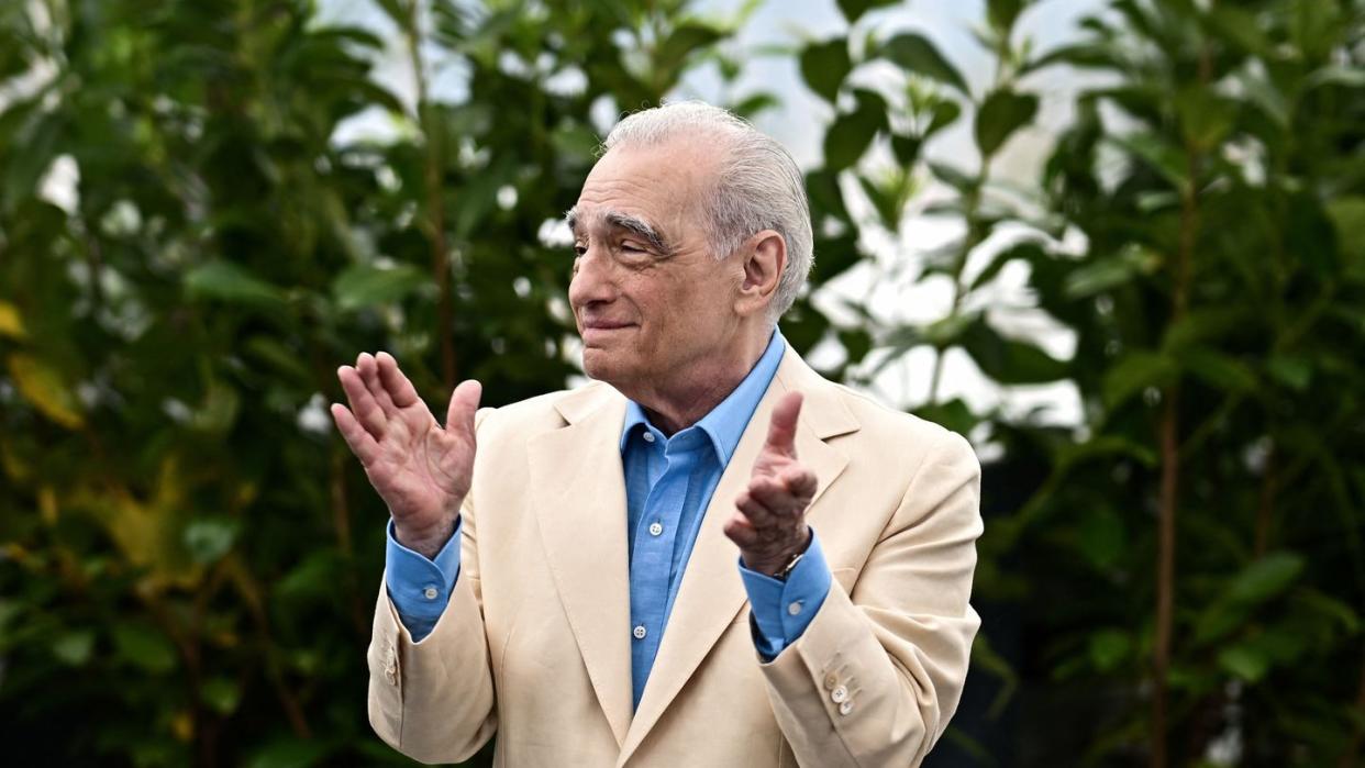 martin scorsese claps his hands while standing in front of greenery, he looks to the left and wears a tan suit jacket over a blue collared shirt