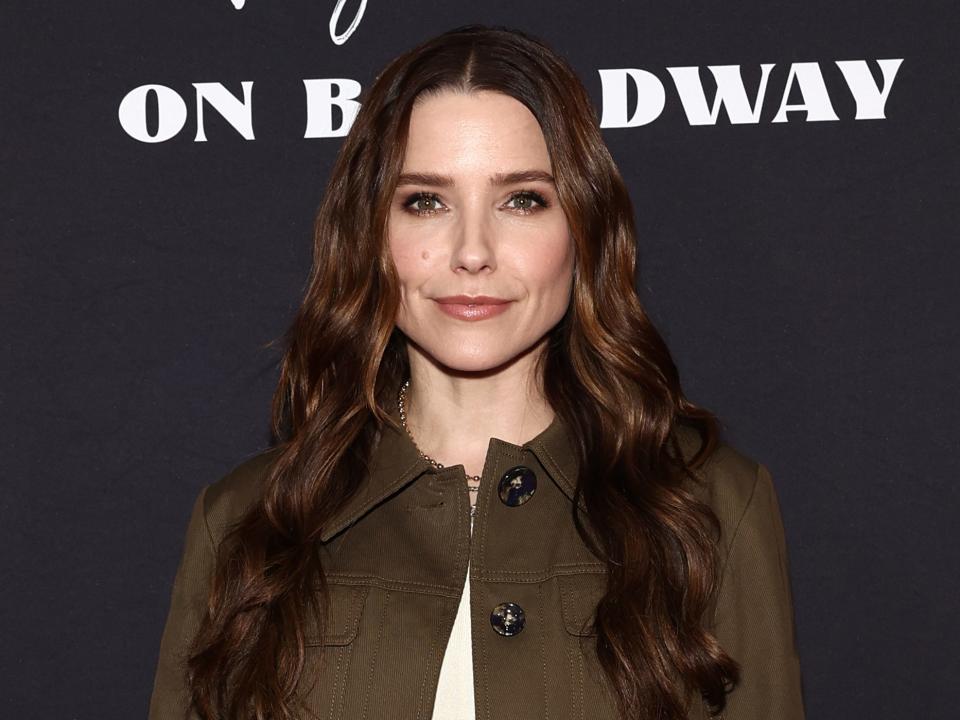 sophia bush in a matching, brown jacket and skirt combo with a white shirt, her hair worn in loose waves, posing on a red carpet