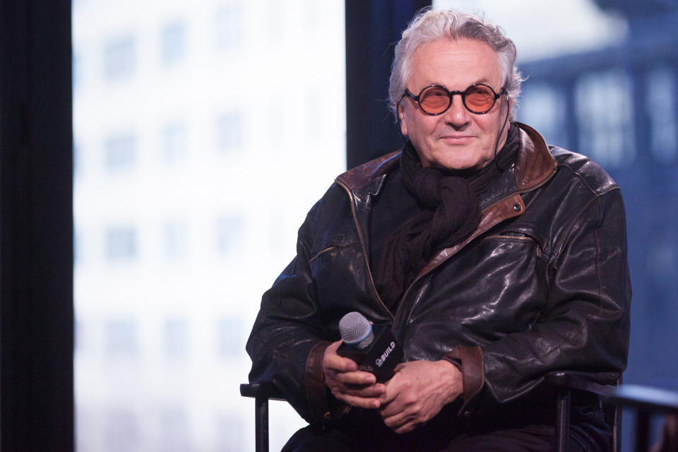 George Miller sits holding a microphone, wearing a leather jacket and scarf