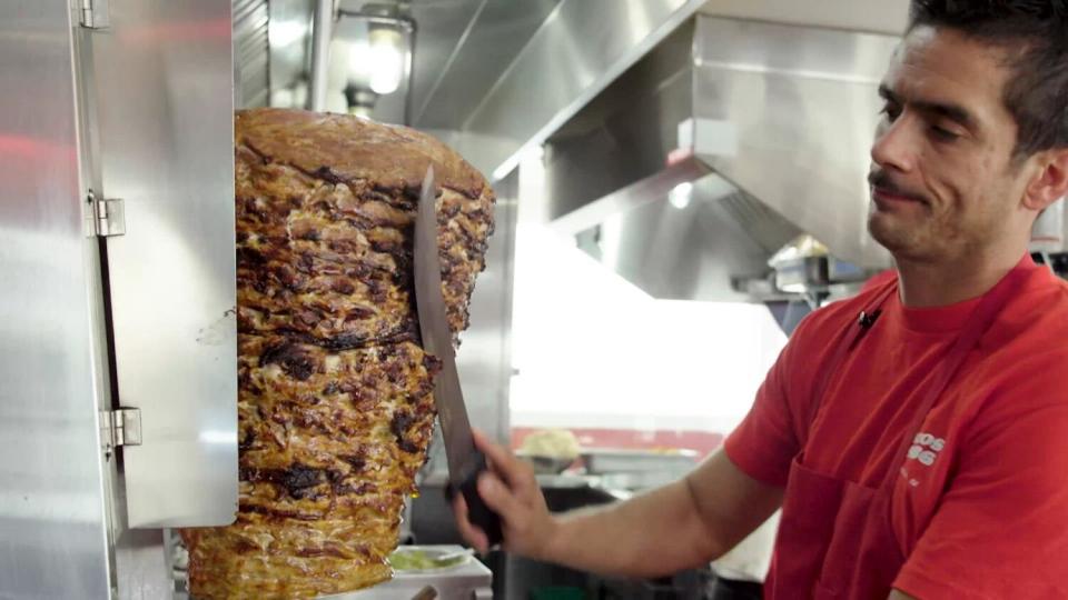 A man stands in a kitchen shaving meat from a spit with a large knife.