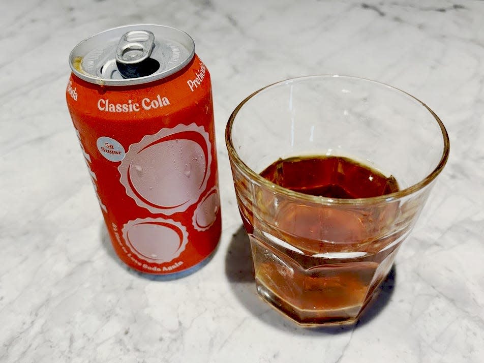 An open can of classic-cola Poppi next to a small, clear glass with brown liquid inside. Both are sitting on a countertop.