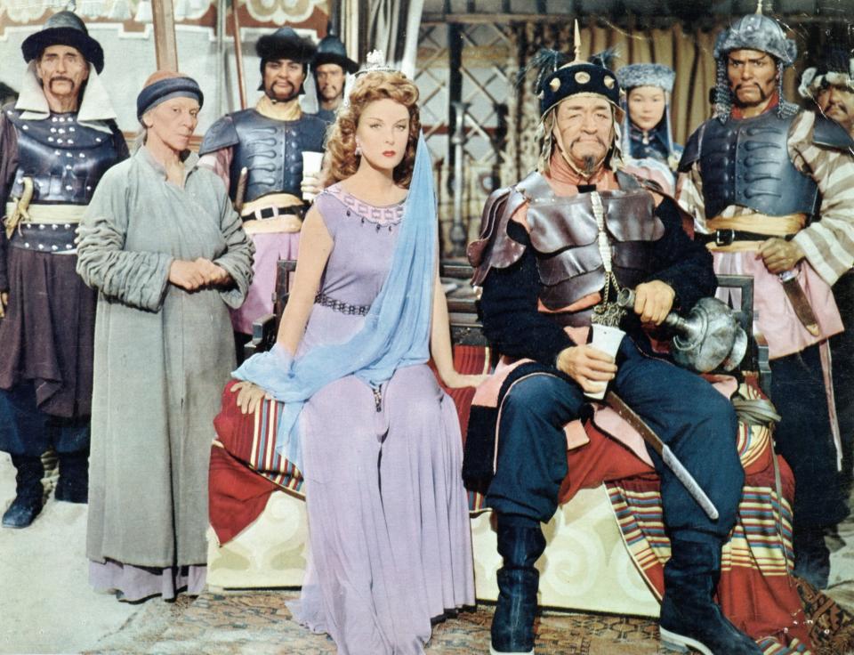 Susan Hayward and John Wayne sitting together surrounded by unidentified actors in a scene from the film 'The Conqueror'