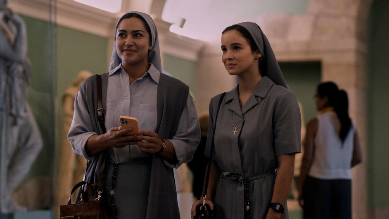  From left to right: Meena Rayann holding a phone while looking up and smiling standing next to Alba Baptista who is also smiling.  