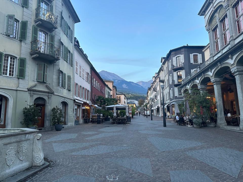 brick-paved pedestrian-only street lined with lamposts and four-story pastel-colored buildings with mountains swiss alps in the distance